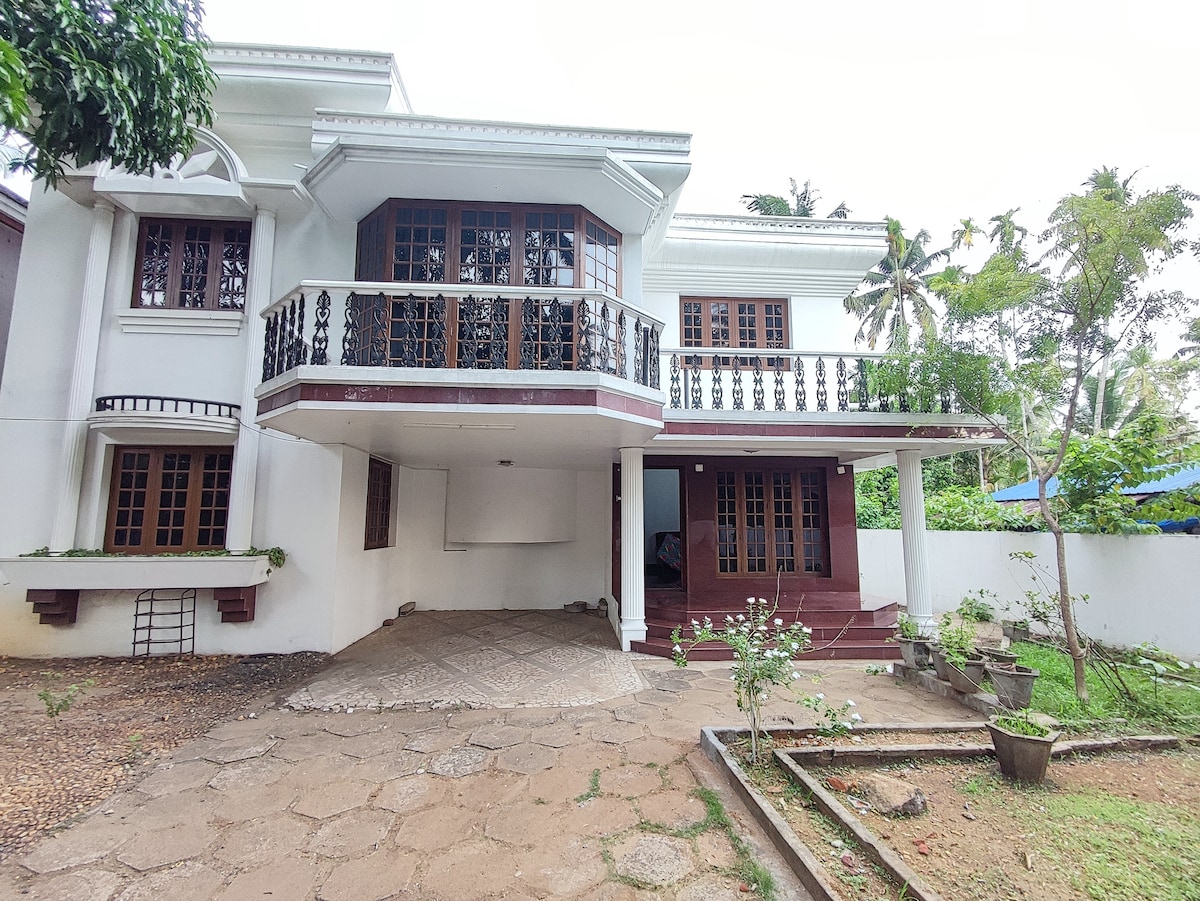 The exterior of a two-story house is presented, featuring a balcony with decorative railings. A stone pathway leads to the entrance, surrounded by a manicured garden with potted plants. Lush greenery is visible in the background, enhancing the inviting setting.