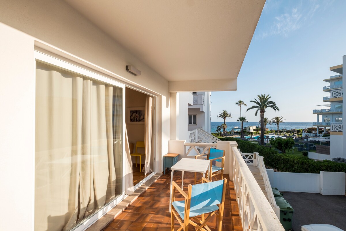 A spacious balcony features a small table and two blue chairs, inviting relaxation. Sheer curtains frame the glass doors, allowing soft natural light to enter. In the background, palm trees gently sway, and the ocean glistens under clear skies.