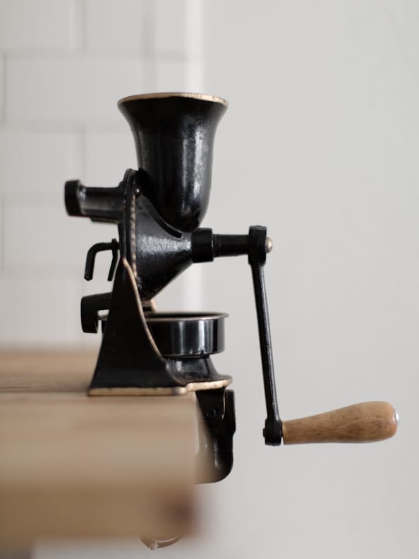 A vintage hand-cranked coffee grinder is displayed on a wooden surface. The grinder features a black and metallic finish with a wooden handle, highlighting its classic design. The background consists of light-colored tiles, adding a subtle contrast.