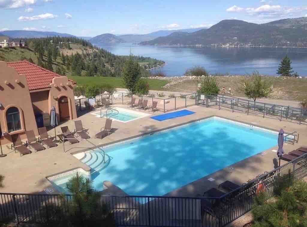 The image captures a serene pool area overlooking a lake, with two swimming pools side by side, surrounded by lounge chairs. Lush green hills and distant mountains create a scenic backdrop, while the clear blue sky reflects on the water's surface.