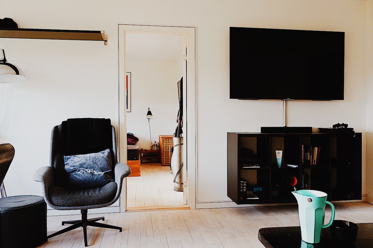A spacious living room features a large television mounted on the wall. A contemporary armchair sits beside a wooden shelving unit filled with books and decor. Light enters through an open doorway leading to another room, creating a sense of openness.