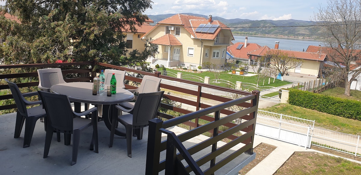 A terrace with a round table and four chairs is visible, providing a scenic view of surrounding houses and the landscape. Lush greenery and a pathway lead towards the river, creating an inviting outdoor space for relaxation.