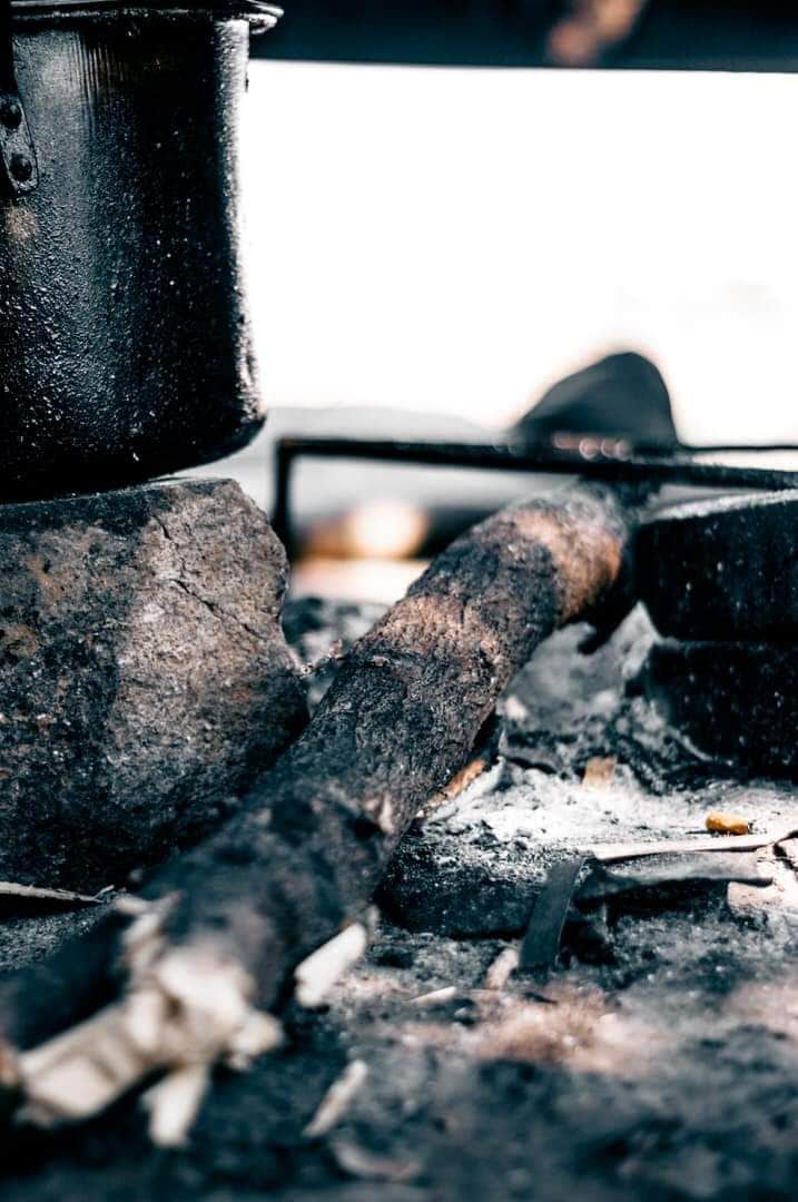 A rustic fire pit is depicted, featuring a cast iron pot resting atop stones. Charred logs lie adjacent to the pit, with ashes scattered on the ground, creating a sense of an inviting cooking area in a natural setting.