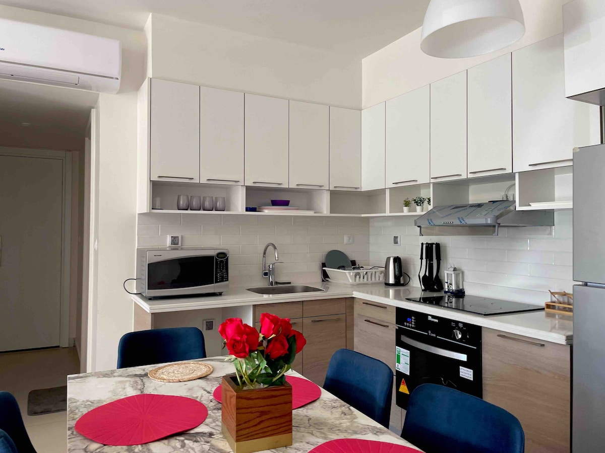 A modern kitchen is depicted, featuring sleek cabinetry and stainless steel appliances. A dining table with blue chairs is positioned in the foreground, adorned with a vase of red roses and placemats. Counter space is utilized for a kettle and a microwave.