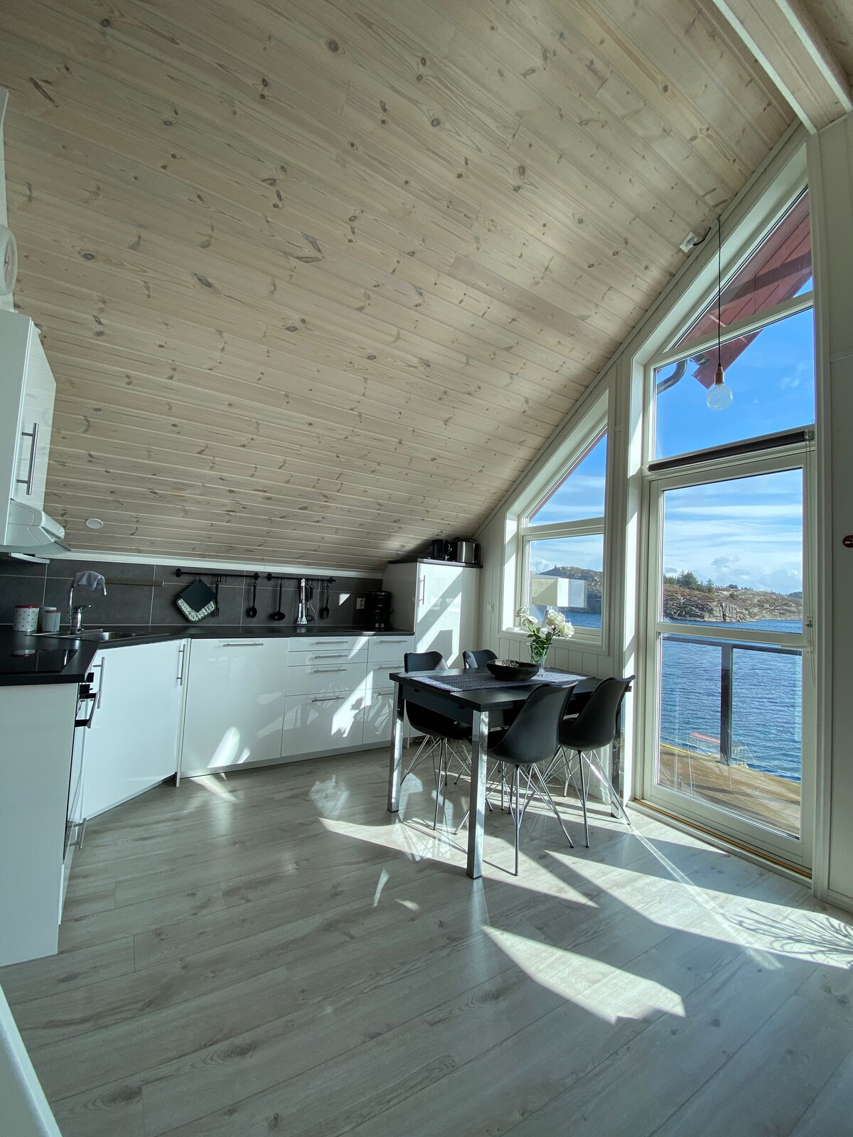 A modern kitchen area features white cabinetry and a black-tiled backsplash. A dining table with four chairs is positioned near large windows, allowing natural light to illuminate the space and offering a view of the water outside.