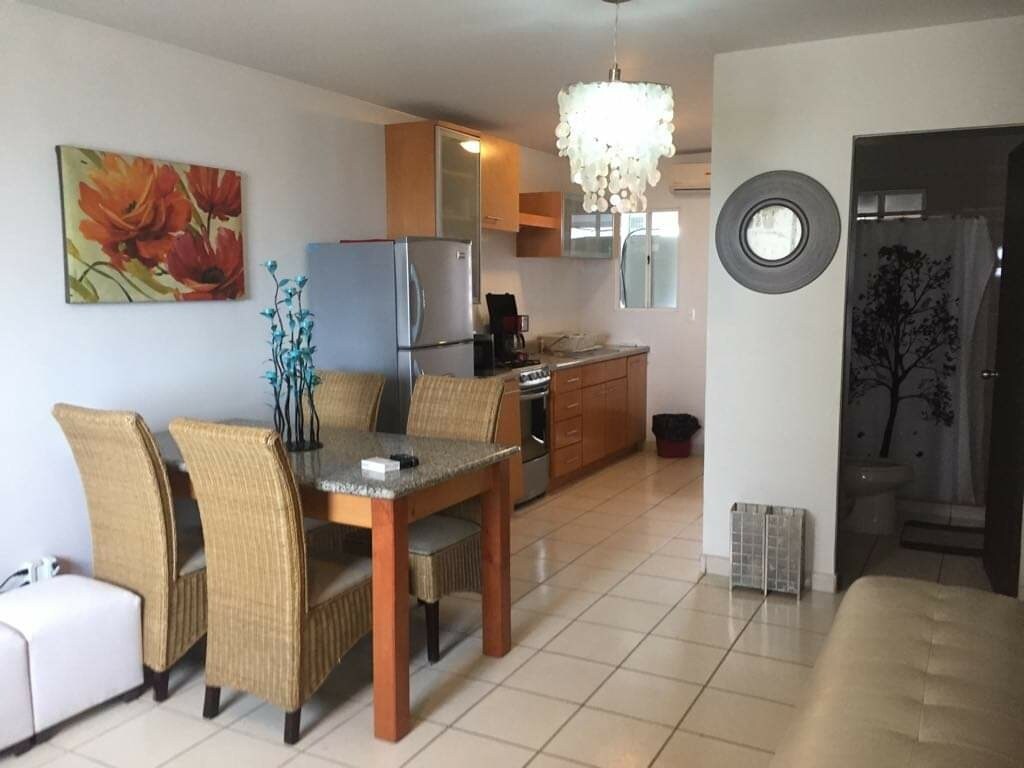 A dining area features a wooden table surrounded by four woven chairs. A modern light fixture hangs from the ceiling. Kitchen appliances, including a refrigerator and stove, are visible along the counter. A colorful floral art piece adds a pop of color to the neutral walls.
