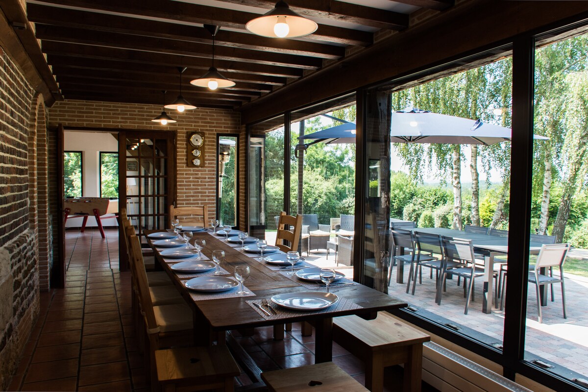 A spacious dining area is displayed with a long wooden table set for multiple guests. Large glass doors provide natural light and views of the outdoor terrace, featuring outdoor seating and umbrellas. Exposed wooden beams and a brick wall add a rustic touch.