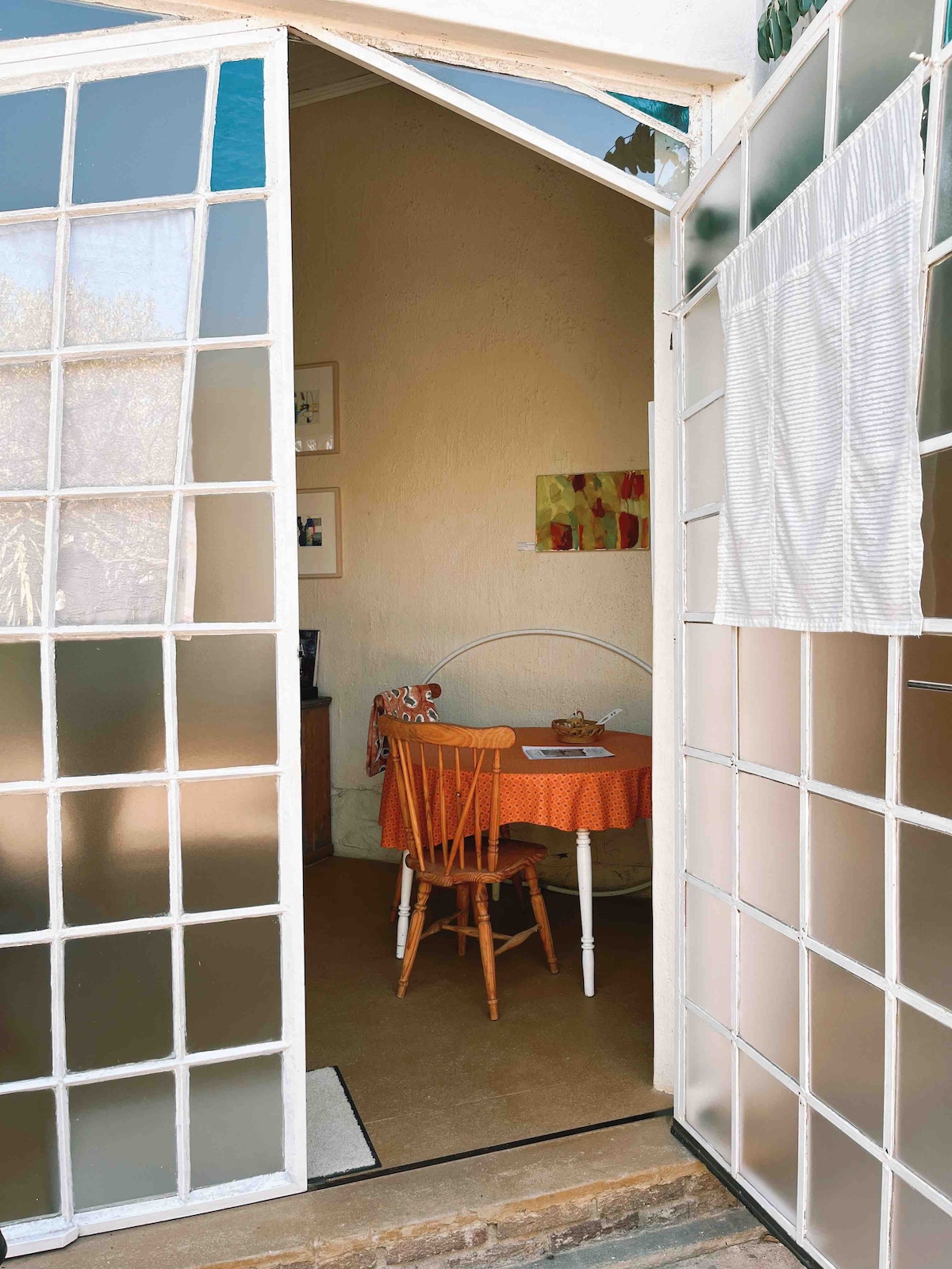 A cozy dining area is visible through glass doors, featuring a small round table covered with an orange tablecloth. A wooden chair with a curved backrest complements the setting. Natural light filters in softly, enhancing the simplicity of the space.