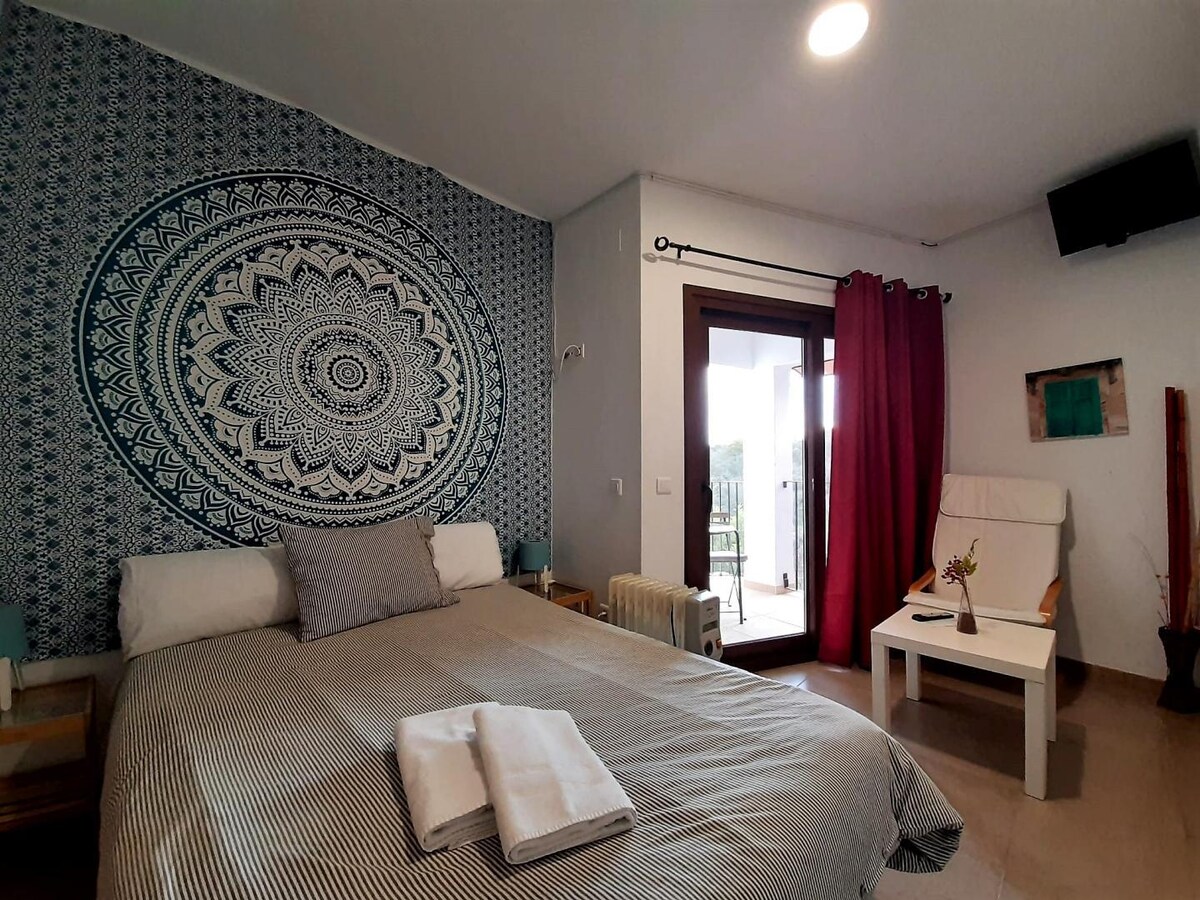 A comfortable sleeping area is highlighted by a decorative mandala wall. The spacious bed is dressed in neutral tones with white towels neatly placed on top. A cozy armchair sits adjacent to a glass door that opens to a balcony, allowing natural light to brighten the room.