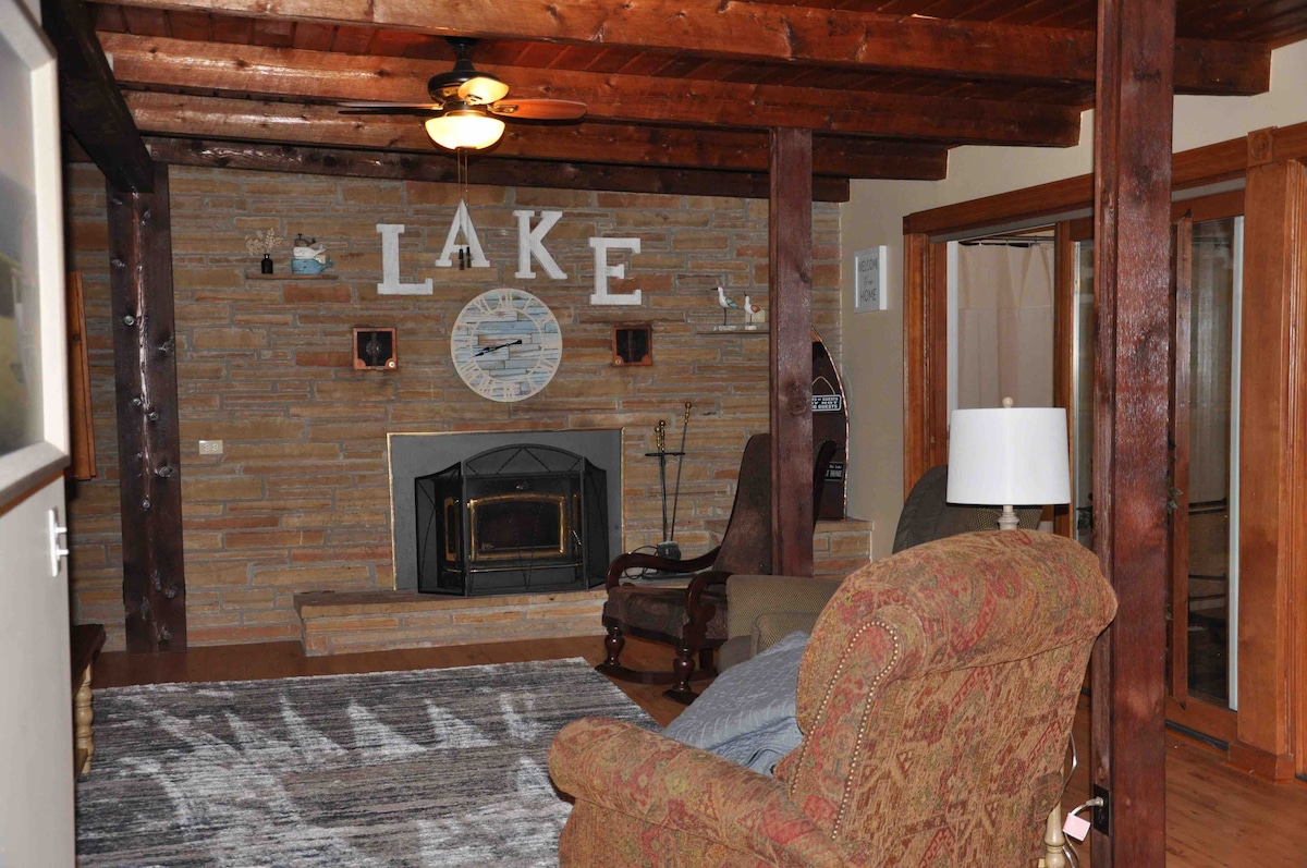A welcoming living area features wooden beams and a stone accent wall with the word 'LAKE' displayed prominently. A comfortable chair and a lamp are positioned beside a cozy fireplace, while a large area rug adds warmth to the wood flooring.