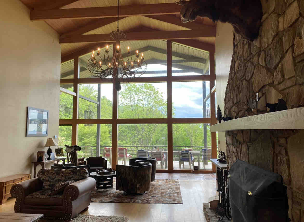 A spacious living area features large windows offering views of the surrounding nature. The room is appointed with comfortable seating, including a leather sofa and unique barrel chairs. A stone fireplace adds character, while a chandelier hangs gracefully from the vaulted ceiling.