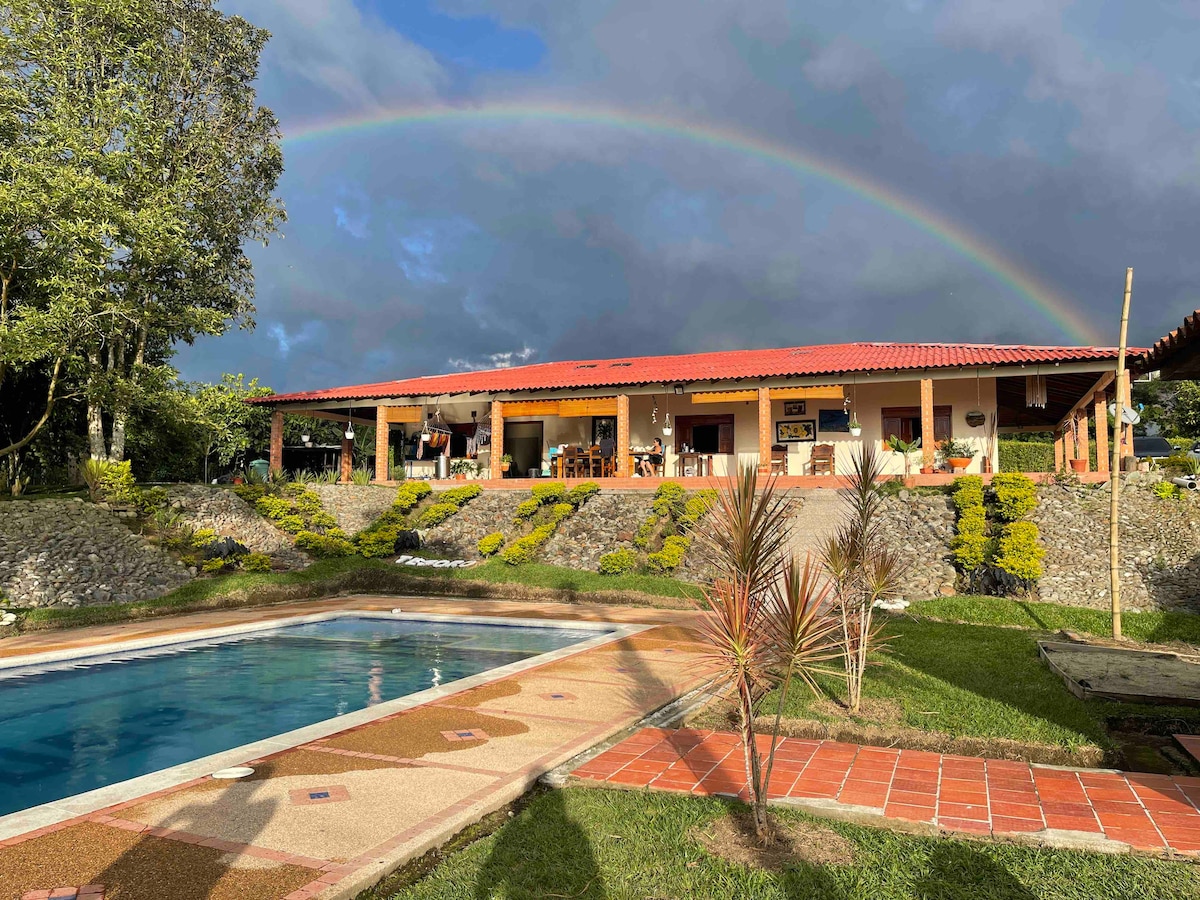A spacious chalet is set against a backdrop of lush greenery under a rainbow. In front, a rectangular pool is framed by manicured gardens, while the building features multiple outdoor seating areas and a traditional red-tiled roof.