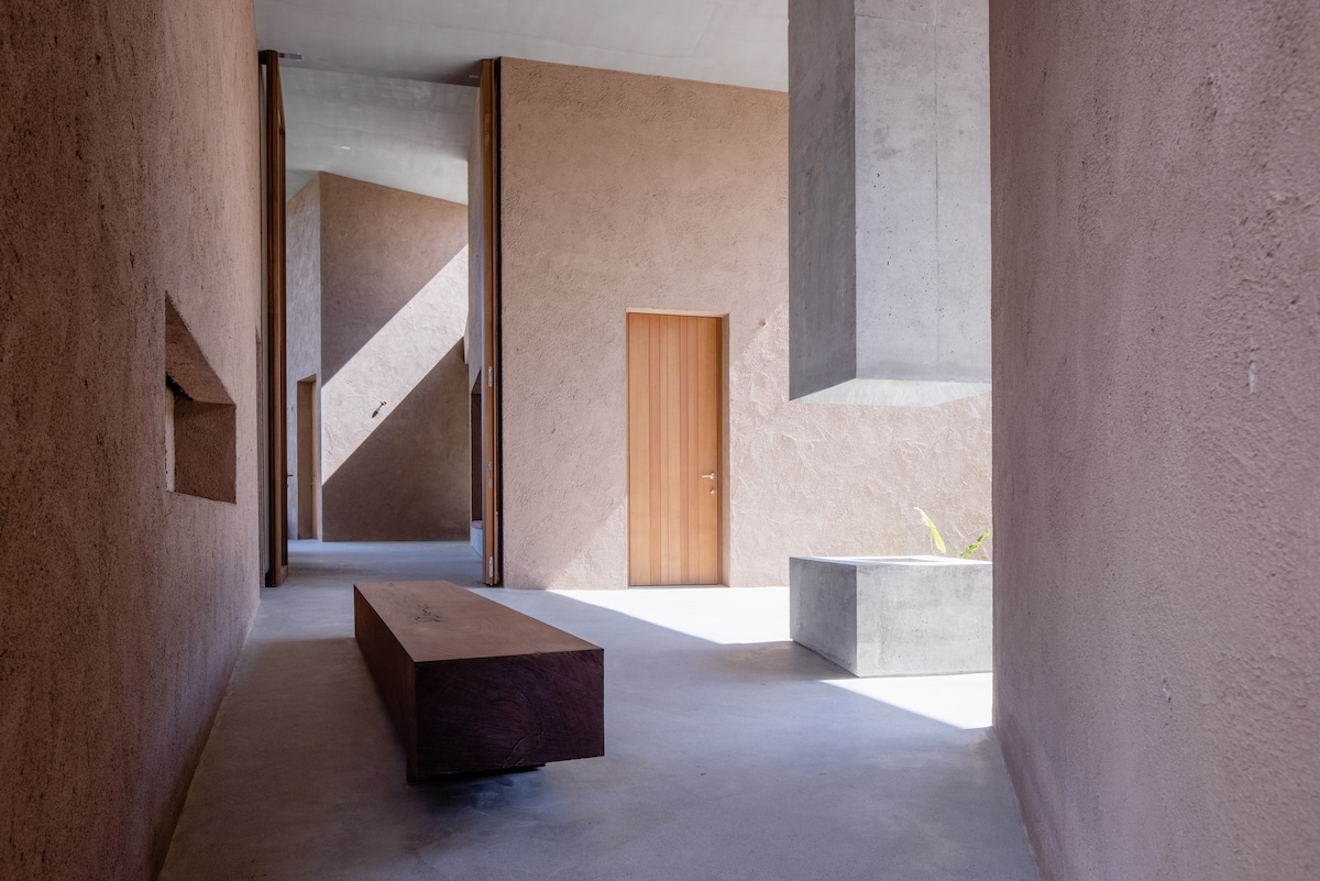 A spacious interior is illuminated by natural light streaming through large openings. The walls feature a warm, textured finish, with a wooden door on one side. A wooden bench and a concrete block element provide minimalist seating and surfaces within the open layout.