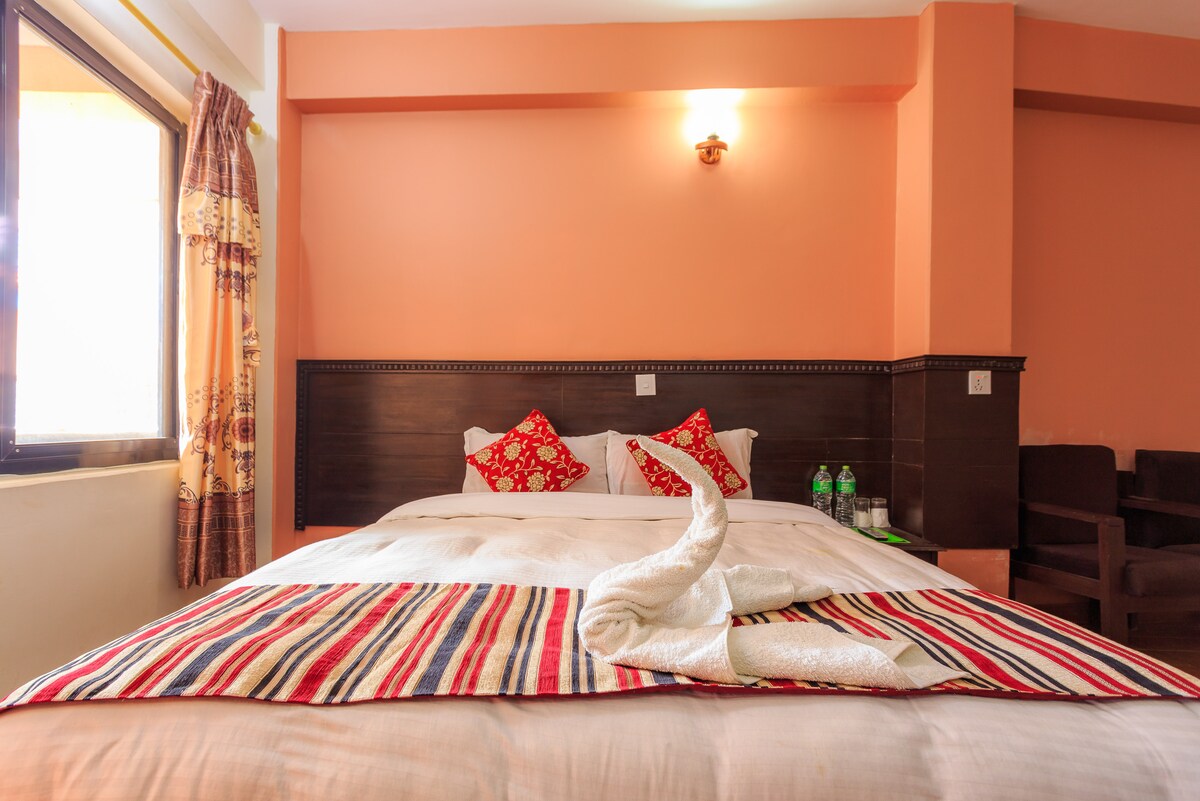 A spacious sleeping area features a neatly made bed adorned with a striped blanket and decorative red pillows. Soft curtains frame the window, allowing natural light to filter through. Two chairs are positioned beside a table, creating a cozy spot for relaxation.