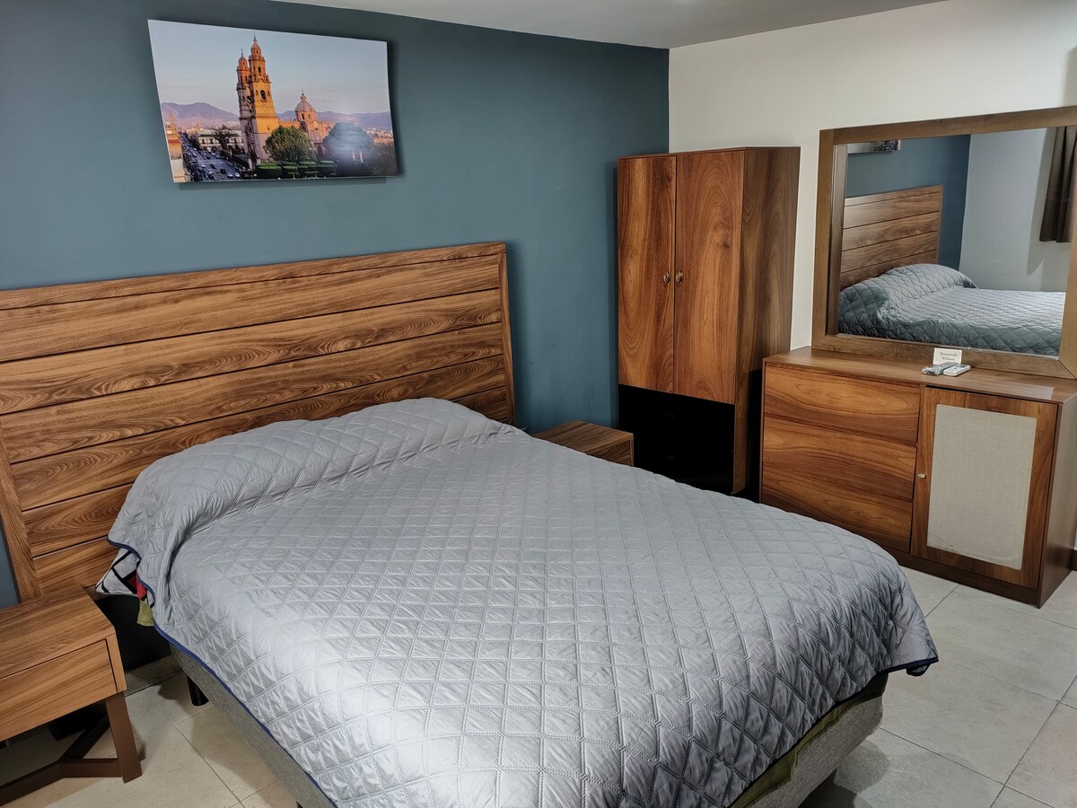A cozy bedroom features a wooden bed frame with a quilted grey coverlet. A flat-screen television is mounted above the bed, displaying a scenic view. A wooden wardrobe and dresser are positioned against the wall, enhancing the room's functionality.