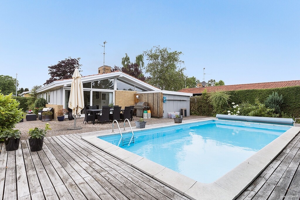 A spacious outdoor area features a clear swimming pool surrounded by a wooden deck. Lounge chairs and a dining table are visible, complemented by potted plants. The house's modern architecture is noted, with large windows allowing for natural light and a serene atmosphere.