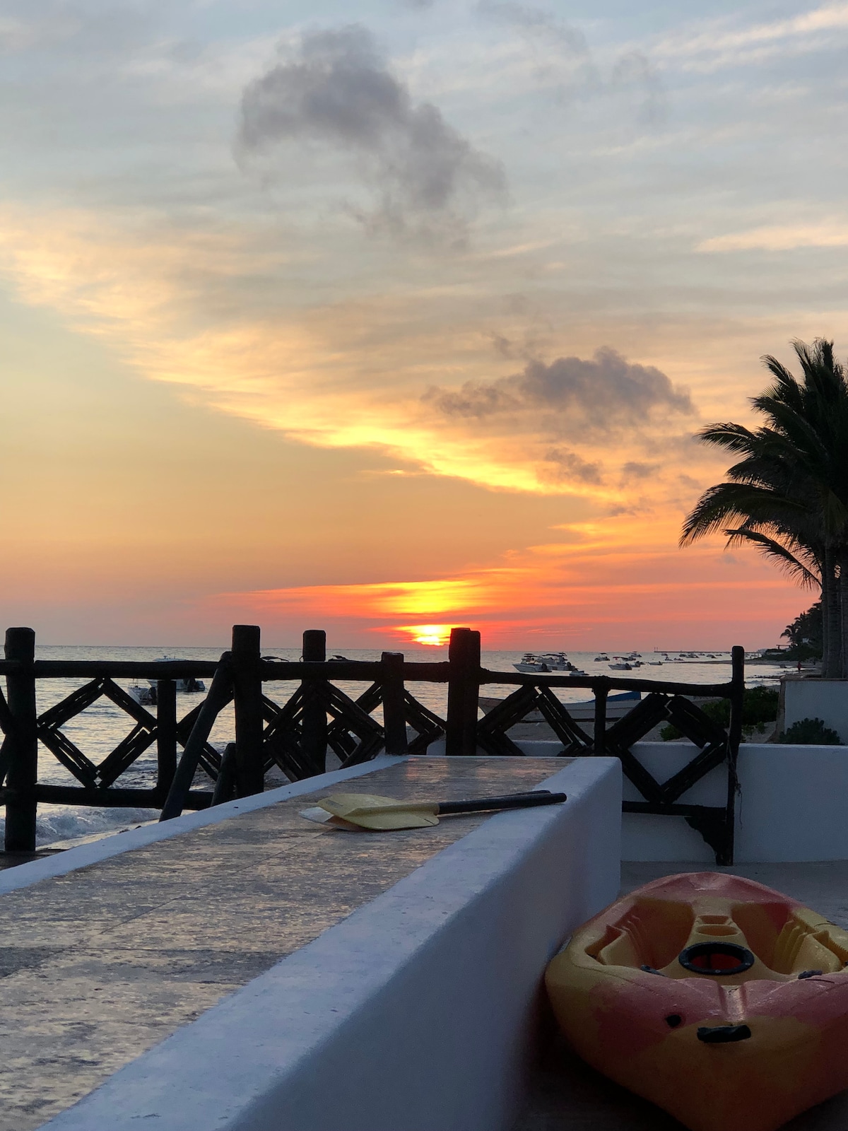 A vibrant sunset casts warm hues across the sky, reflecting on the ocean's surface. A wooden fence outlines the property, and a kayak rests nearby on a smooth white surface. Palm trees frame the scene, enhancing the coastal atmosphere.