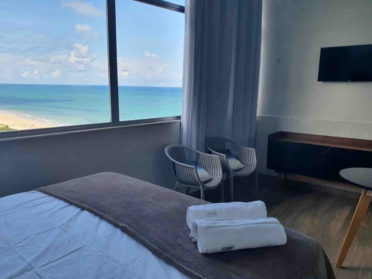 A spacious room is featured with a large window offering a clear view of the sea. Two chairs are positioned beside the window, allowing for relaxation while looking out at the beach. Fresh towels are neatly arranged on the bed.