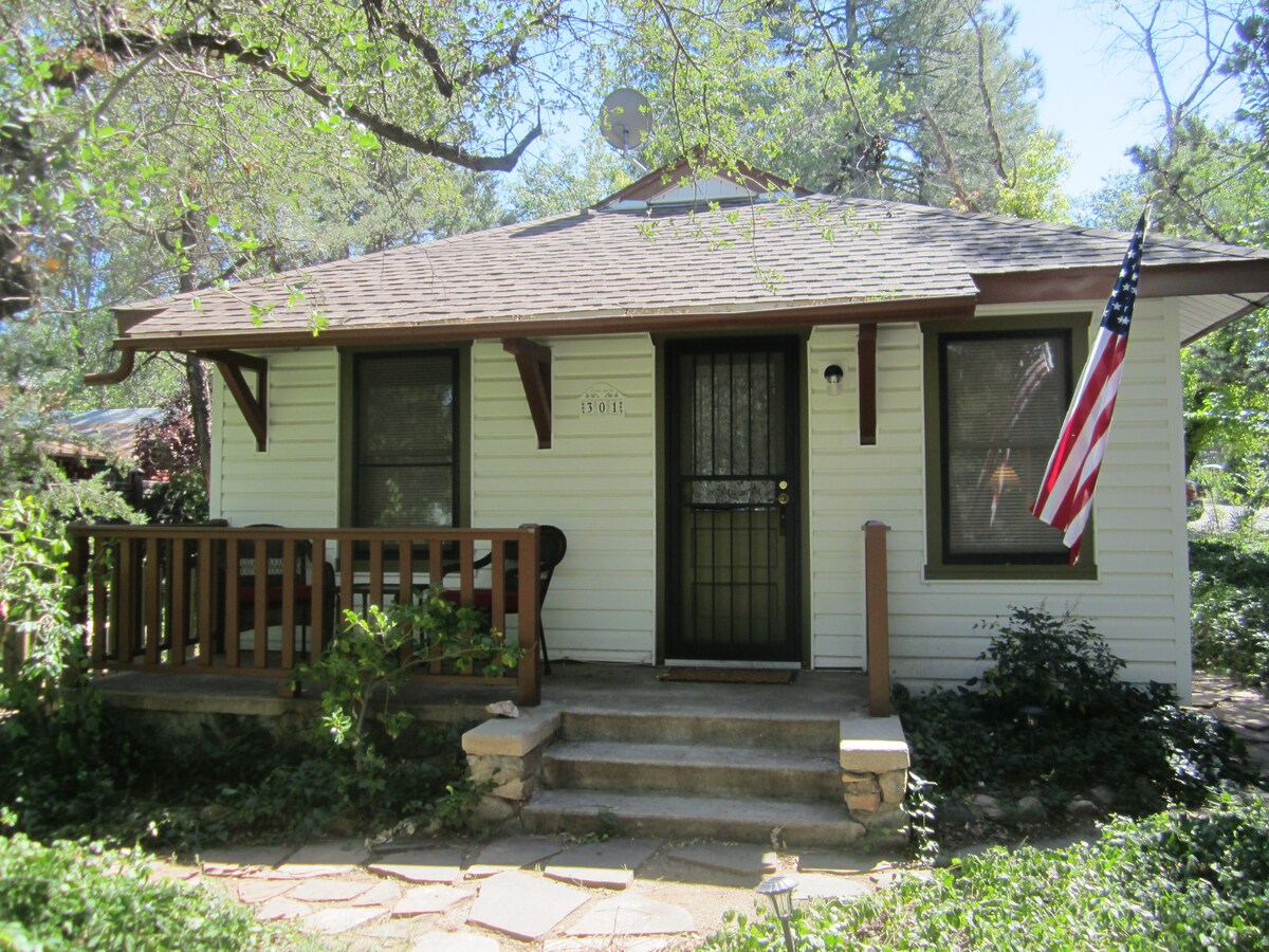 A charming cottage exterior is featured, surrounded by greenery and mature trees. A small front porch with wooden railings is visible, and a traditional American flag is displayed by the front door. Stone steps lead to the entrance, enhancing the inviting appearance.