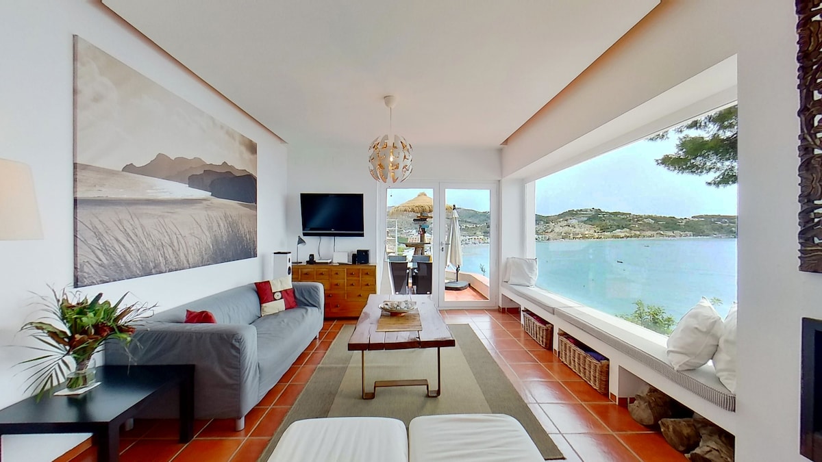 A modern living area features a spacious gray sofa and a central wooden coffee table, with panoramic windows offering unobstructed views of the sea and mountains. Natural light brightens the space, complemented by a television and decorative elements, creating an inviting atmosphere.