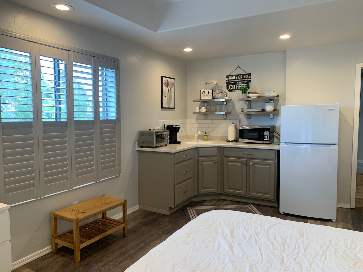 A well-equipped kitchenette is displayed, featuring a refrigerator, microwave, and coffee maker. Shuttered windows allow natural light to filter in. The space includes gray cabinetry and a small wooden bench for convenience, providing a functional area for meal preparation.