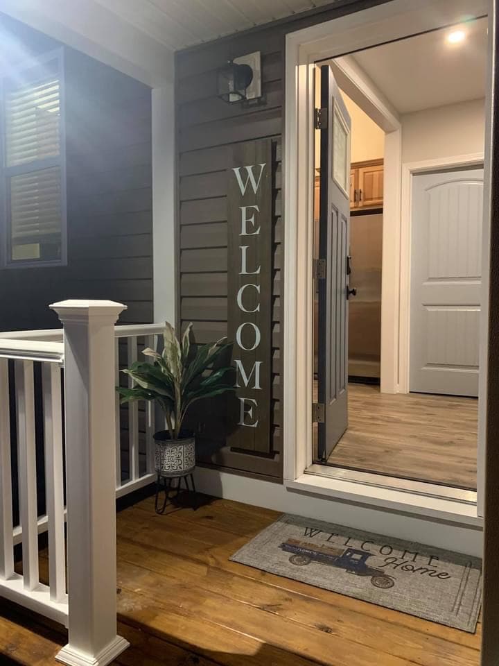 An inviting entrance is showcased with a welcoming sign and a potted plant at the base of a white railing. A doormat with 'WELCOME HOME' is positioned at the threshold, leading to a view of the interior doorway illuminated by warm light.
