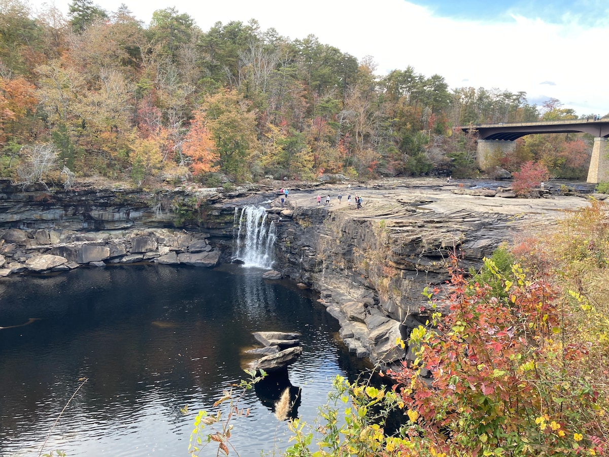 A serene landscape features a waterfall cascading into a tranquil pool, surrounded by rocky outcrops. Colorful autumn foliage adds warmth to the scene. A bridge arches overhead, integrating natural beauty with structural design.