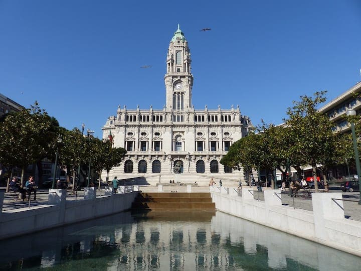 The image showcases a grand historic building with an ornate façade and a tall clock tower, set against a clear blue sky. A reflecting pool is visible in the foreground, with neatly trimmed trees lining the square, adding to the serene ambiance.
