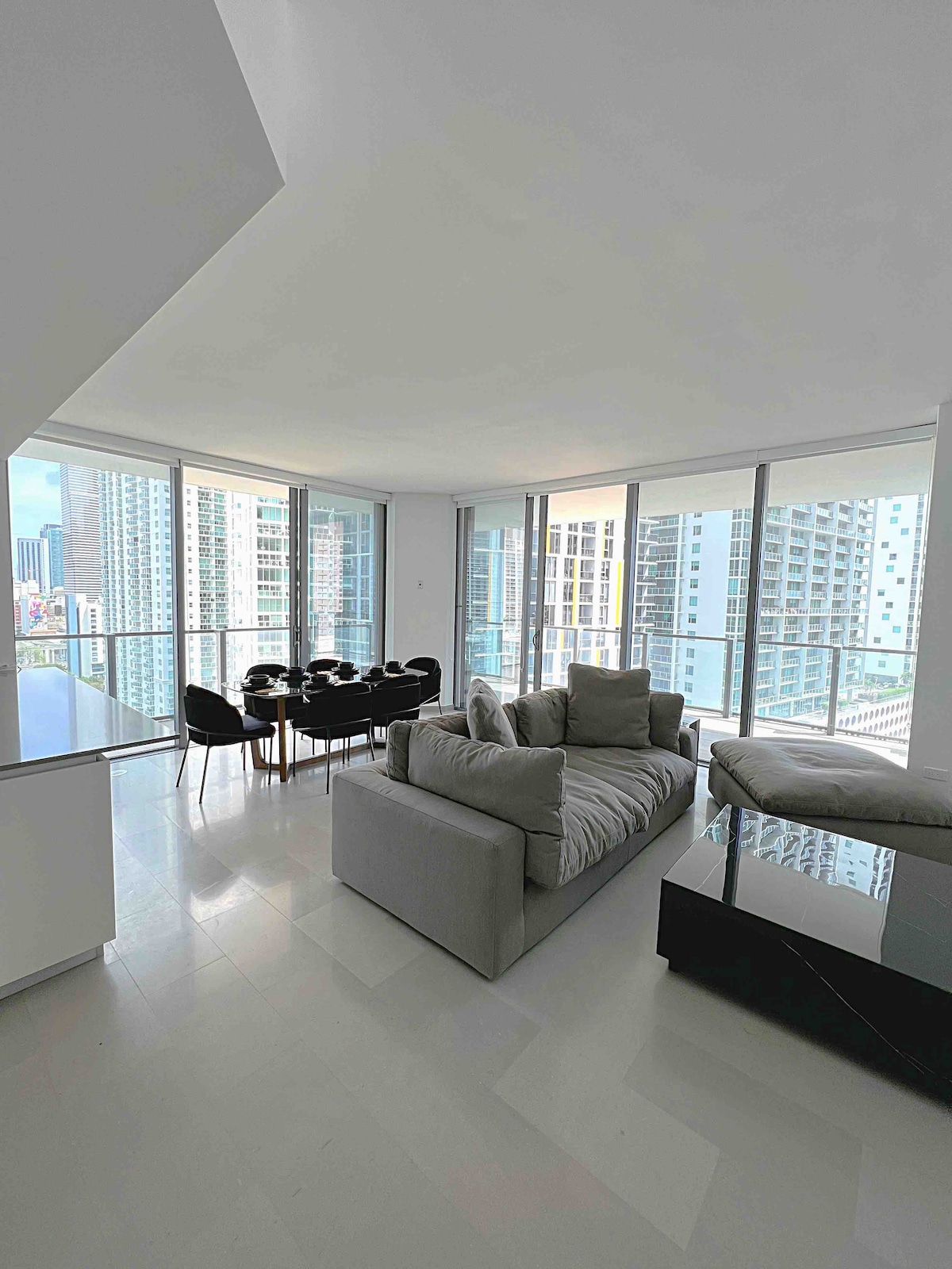 A modern living area is presented with large floor-to-ceiling windows, allowing natural light and showcasing city views. An oversized light grey couch with plush cushions is positioned beside a sleek glass table. A formal dining setup for six is visible in the background.
