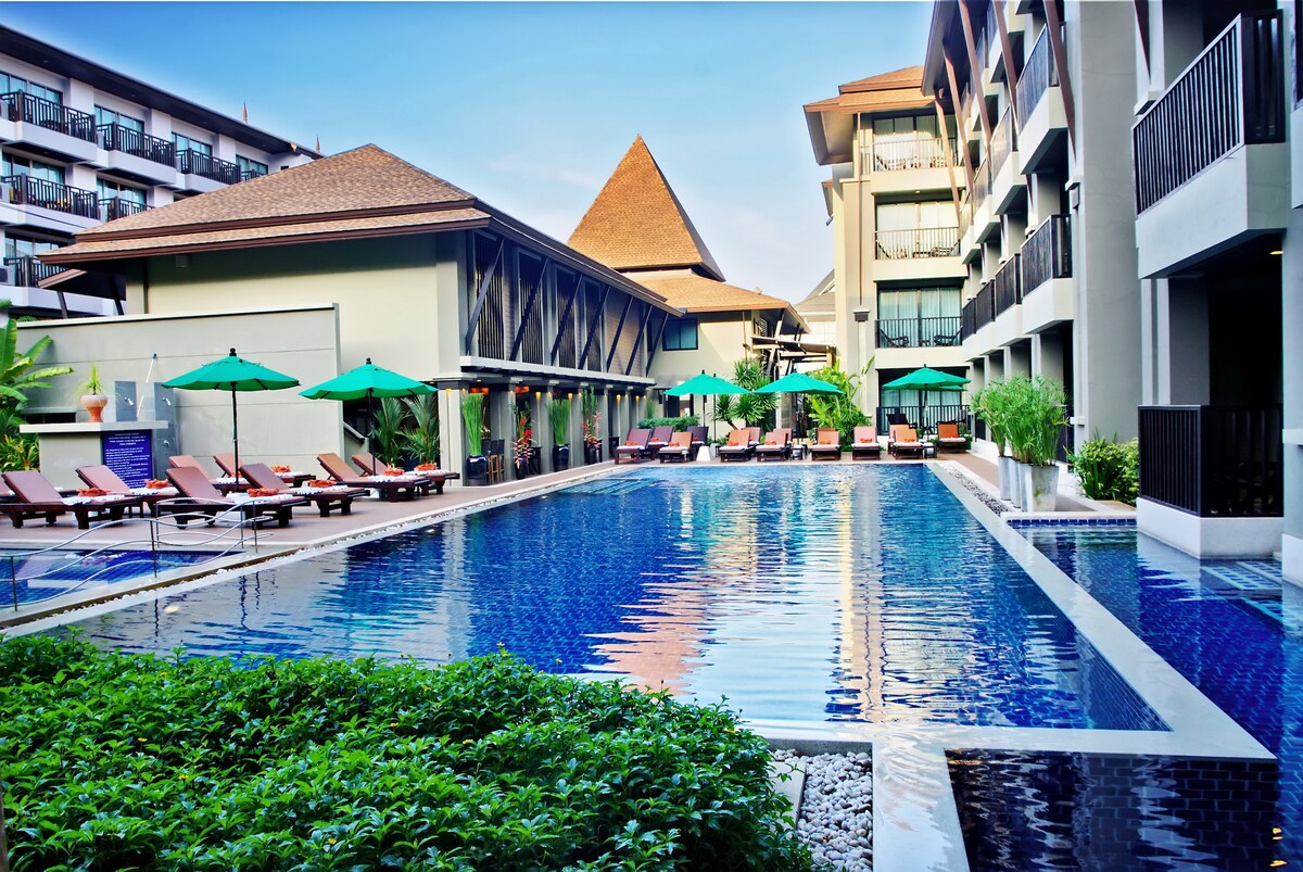 An expansive swimming pool is surrounded by comfortable lounge chairs, shaded by green umbrellas. The sleek water reflects the clear sky and the surrounding architecture, which features traditional and contemporary design elements. Lush greenery enhances the serene atmosphere of the pool area.