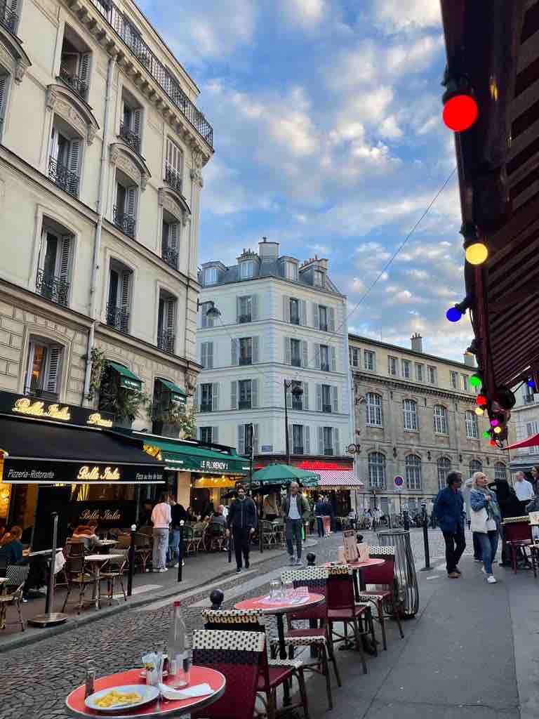 An inviting street scene in Montmartre, featuring outdoor dining tables with red chairs and a cobblestone path. Restaurants line both sides of the street, offering shaded seating beneath awnings. The sky above is a mix of soft clouds, enhancing the lively atmosphere of the neighborhood.