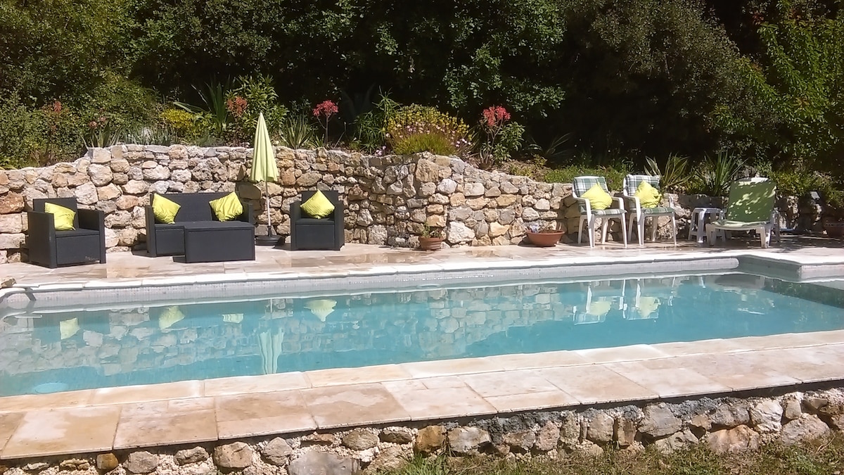A private pool area is bordered by natural stone walls, adorned with vibrant plants. Stylish seating arrangements are positioned nearby, featuring comfortable chairs with bright cushions. An inviting yellow umbrella offers shade, complementing the tranquil setting for relaxation.