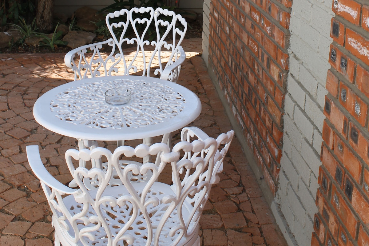 A quaint outdoor seating area is presented with two white, intricately designed chairs and a matching round table. The table features a central spot for an ashtray, while the brick wall and patio stones create a rustic backdrop.