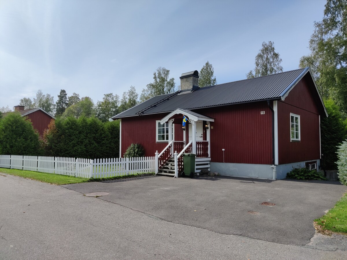 A charming red house with a gray roof features a welcoming entrance with steps leading to the front door. A white picket fence outlines the property, while greenery lines the front, providing a sense of privacy. The street appears quiet, surrounded by residential woods.