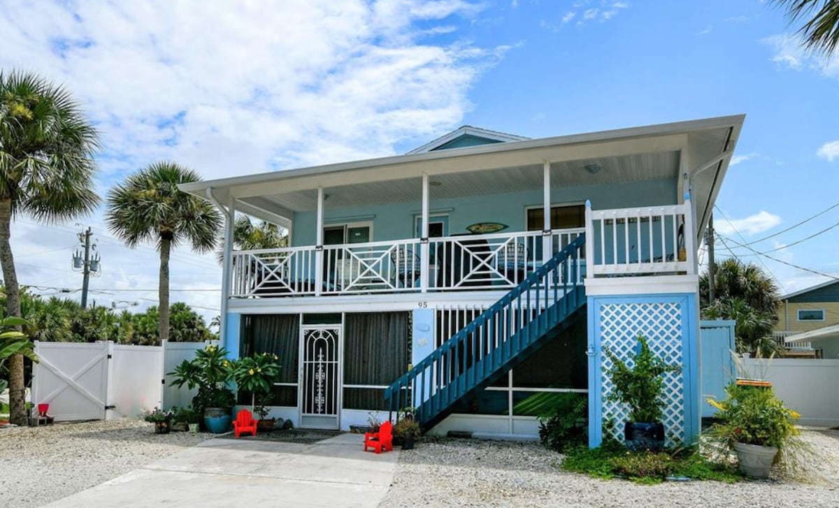 The exterior of the house is showcased, featuring a two-story structure with a blue façade. A staircase leads to the upper level, which is adorned with a porch that includes white railings and welcoming outdoor seating. Palms and other greenery surround the property, complementing the coastal ambiance.