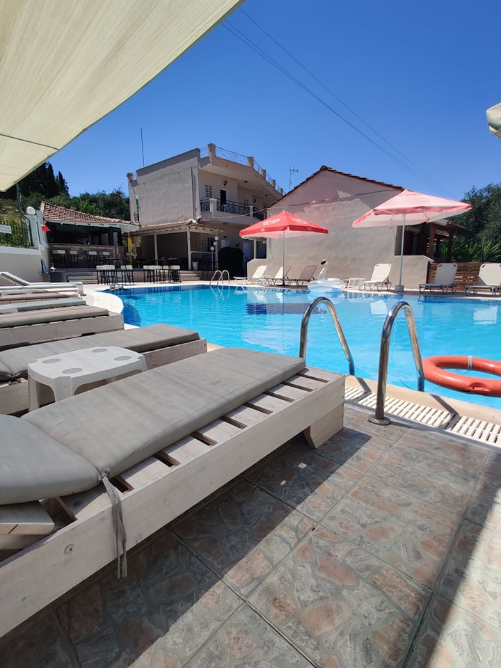 A clear swimming pool surrounded by light lounge chairs and shaded by umbrellas. Sunlight reflects off the water's surface, while a wooden deck leads to the pool entrance. Nearby, a bar area and a building can be seen, creating a relaxed outdoor environment.