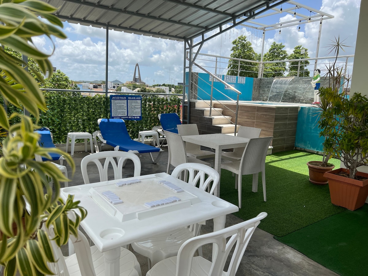 An outdoor seating area features white tables and chairs alongside loungers. A small green lawn and potted plants are visible, creating a refreshing environment. A glimpse of a pool area can be seen in the background, with a distant view of a landmark structure.