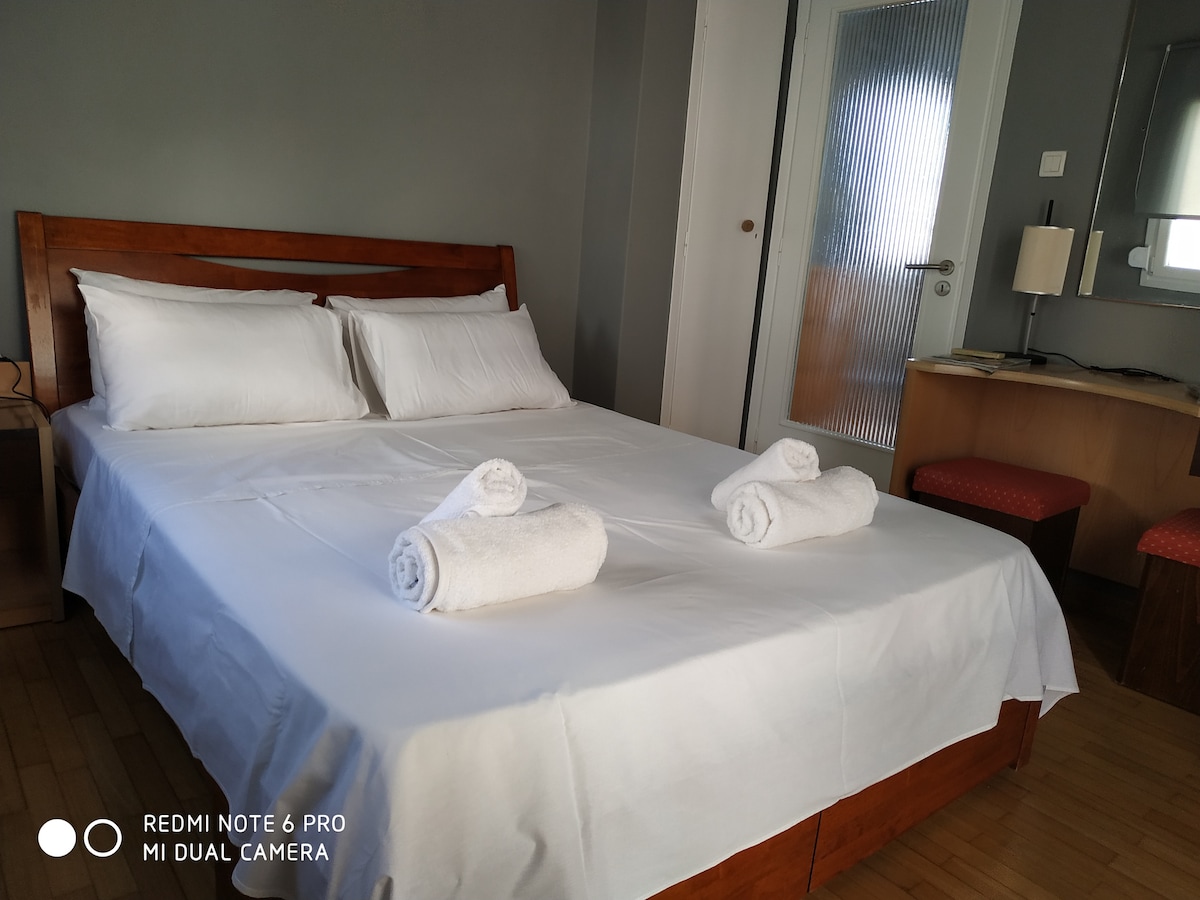 A well-lit bedroom is presented, featuring a spacious bed with crisp white linens. Two rolled towels are neatly arranged on the bed. A window allows for natural light, and a small desk with a lamp and chair is positioned nearby, enhancing the functionality of the space.