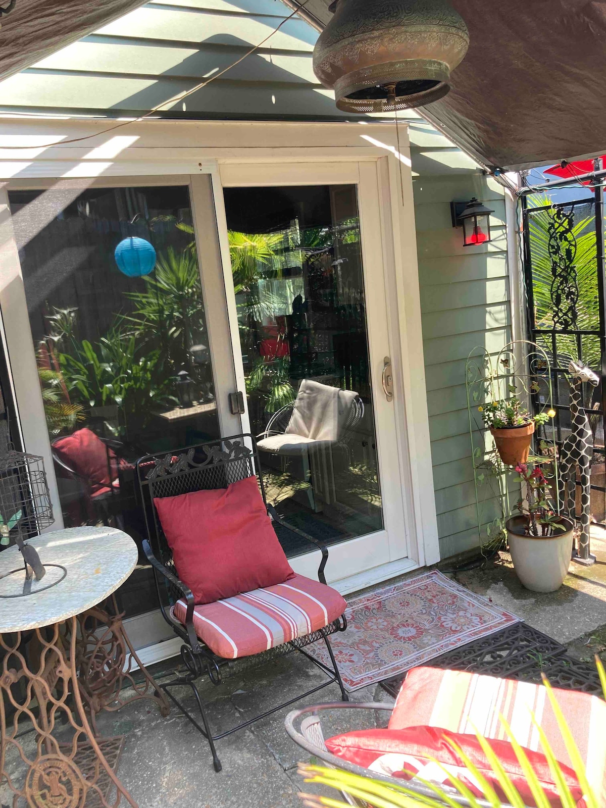 A cozy patio area is visible, featuring a small round table and a comfortable chair with striped cushions. Lush greenery surrounds the space, complemented by a hanging lantern and a vibrant blue globe. A door leads inside, reflecting natural light.