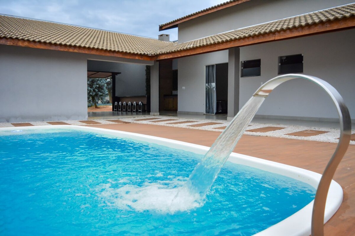 A refreshing pool is featured, with a sleek water feature creating gentle ripples in the clear blue water. The pool area is surrounded by a tiled deck, leading to a modern structure with a textured facade and large glass doors.
