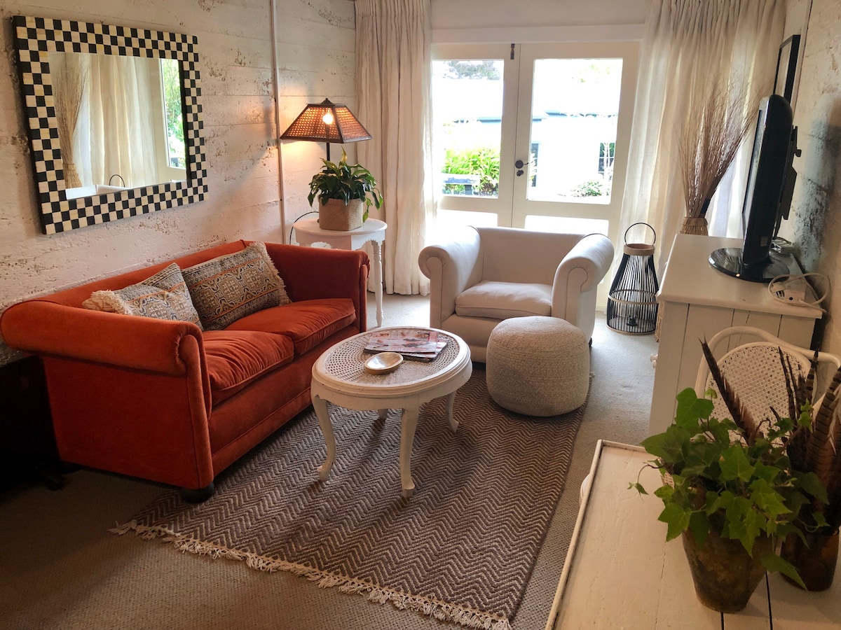 A comfortable living area is furnished with an orange sofa and a white armchair. A round table occupies the center, accompanied by a light-colored ottoman. Natural light enters through large windows dressed with sheer curtains, highlighting the textured walls and the plant on the side table.