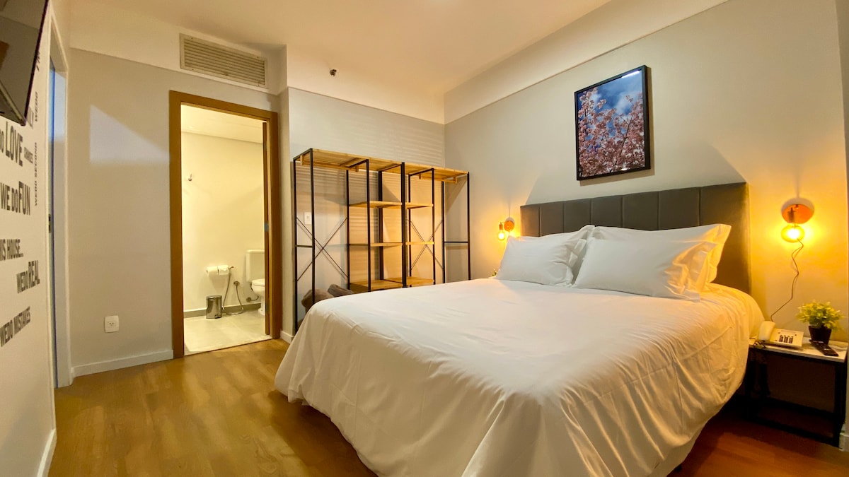 A well-lit bedroom is equipped with a spacious bed covered in crisp white linens. Shelving units are positioned against the wall, while a framed nature print adorns the headboard area. A doorway leads to a bathroom, enhancing the room's practicality.