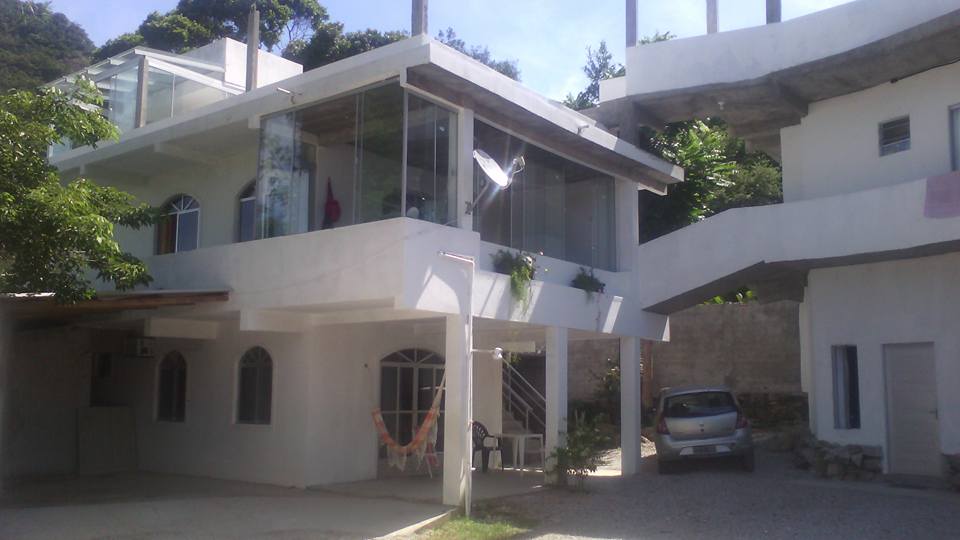 A modern two-story building is shown, featuring large glass windows and an open-air design. The exterior is painted white, with a small car parked nearby. A shaded area with chairs and a hammock enhances the inviting outdoor space.
