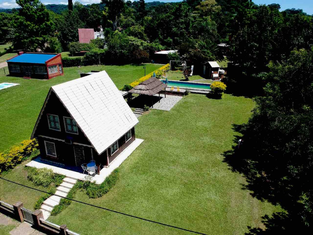 An aerial view of a chocolate-style cabin is shown, surrounded by a spacious green lawn. A large swimming pool, children's play area, and shaded outdoor dining area can be seen, providing ample space for relaxation and activities.