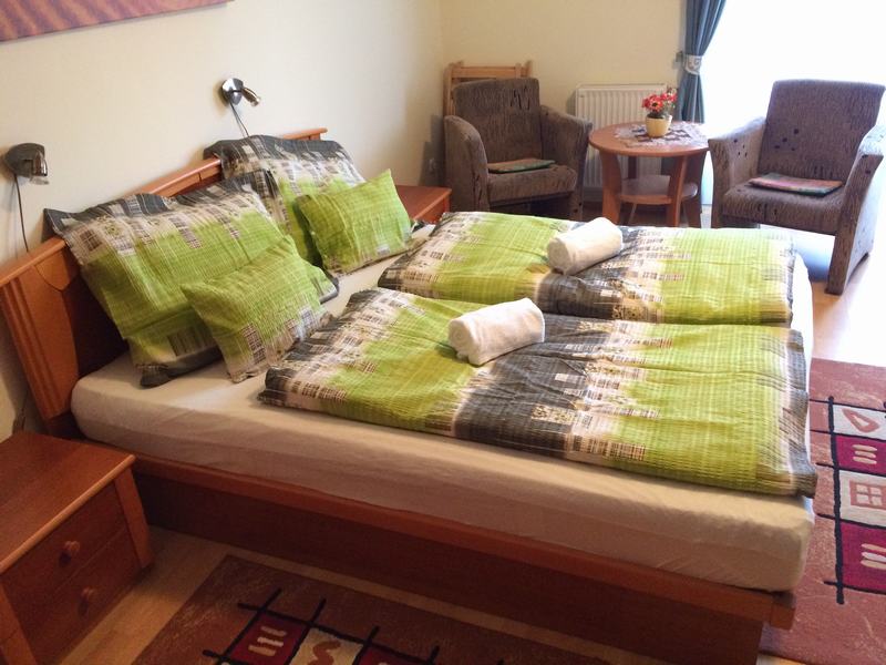A cozy sleeping area features a spacious bed adorned with patterned green bedding and matching pillows. Two white towels are neatly placed on the bed. A small table is set up beside two armchairs, offering a relaxing spot within the room.
