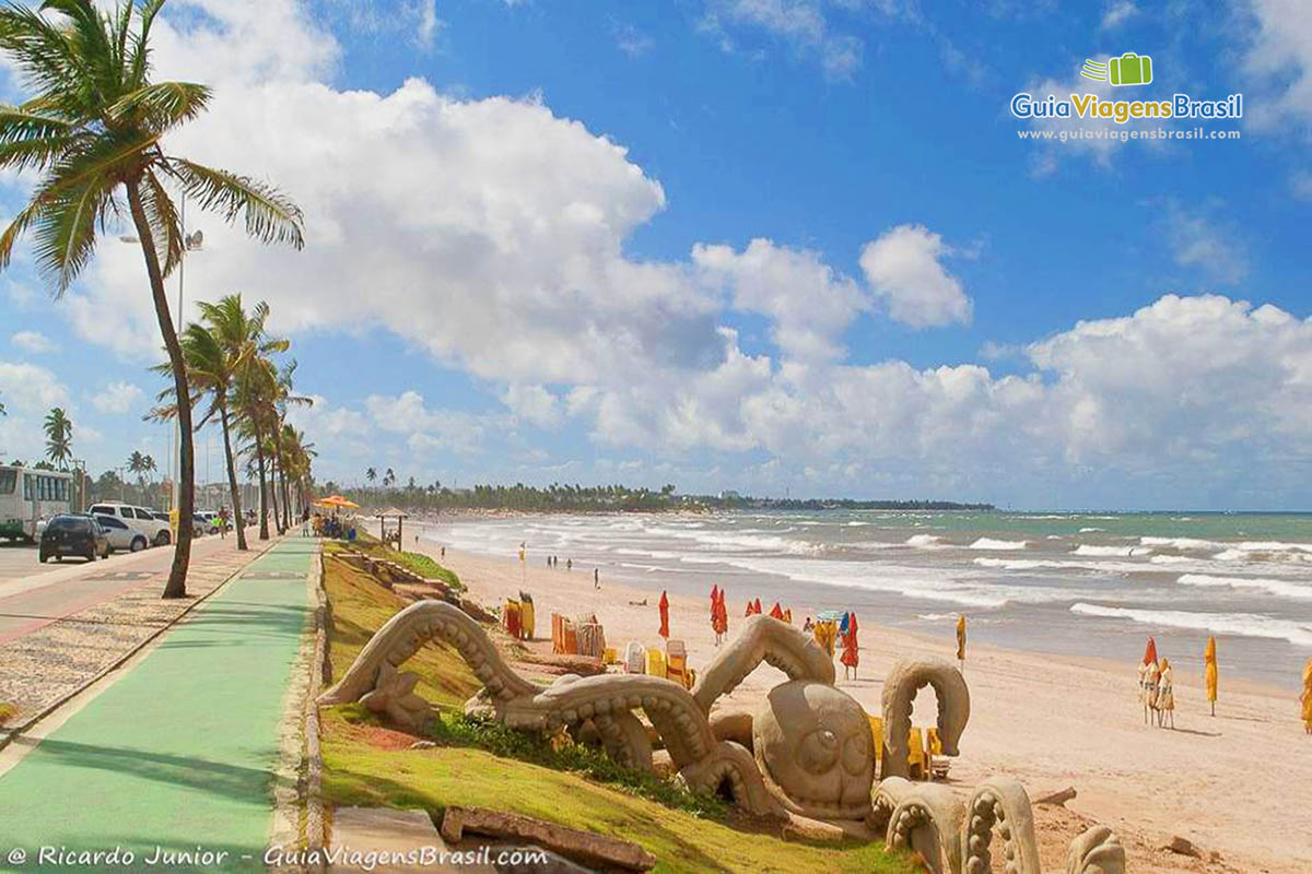 A scenic beach view features a wide shoreline lined with palm trees swaying in the breeze. Sand sculptures, including an octopus, are displayed prominently in the foreground, while beachgoers can be seen enjoying the sun and surf in the distance under a partly cloudy sky.