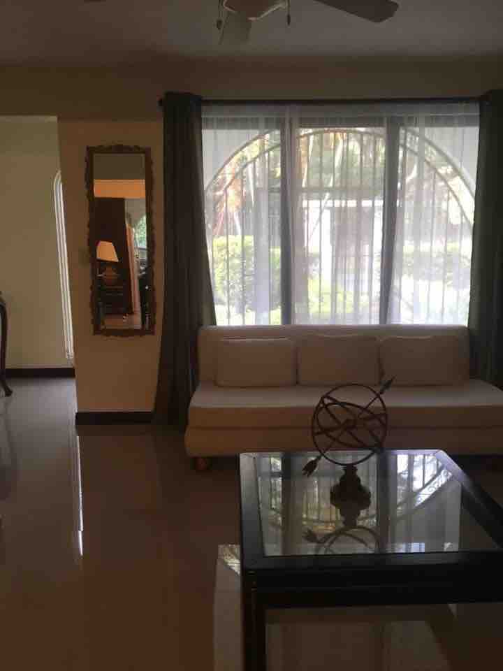 A bright living area features a light-colored sofa facing a glass-top coffee table. Large windows with sheer curtains allow natural light to fill the space, while a decorative mirror enhances the room's openness. A glimpse of greenery is visible through the window.