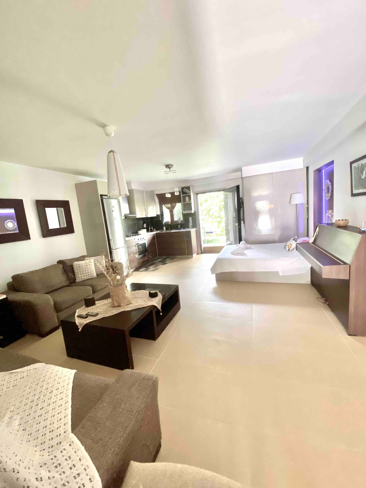 A spacious living area features a comfortable seating arrangement with two sofas and a coffee table. The well-equipped kitchen is visible in the background, with natural light filtering through the sliding glass doors leading to the outdoor space.