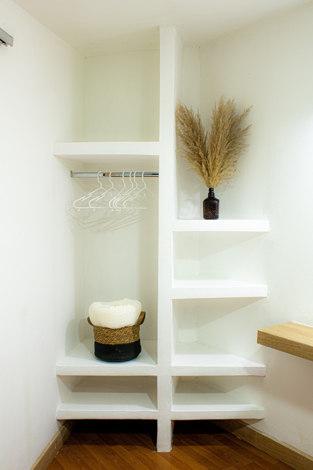 A minimalist closet space is presented with white walls and wooden accents. Several hangers are neatly arranged on a rod, while shelves display a decorative vase and a woven basket, contributing to organized and functional storage options.