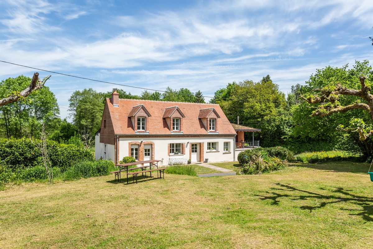 A spacious villa is set against a backdrop of trees and greenery, featuring a welcoming facade with multiple windows and a sloped roof. A large lawn extends in the foreground, with gardening areas and a table positioned for outdoor gatherings. The atmosphere reflects a serene countryside setting.